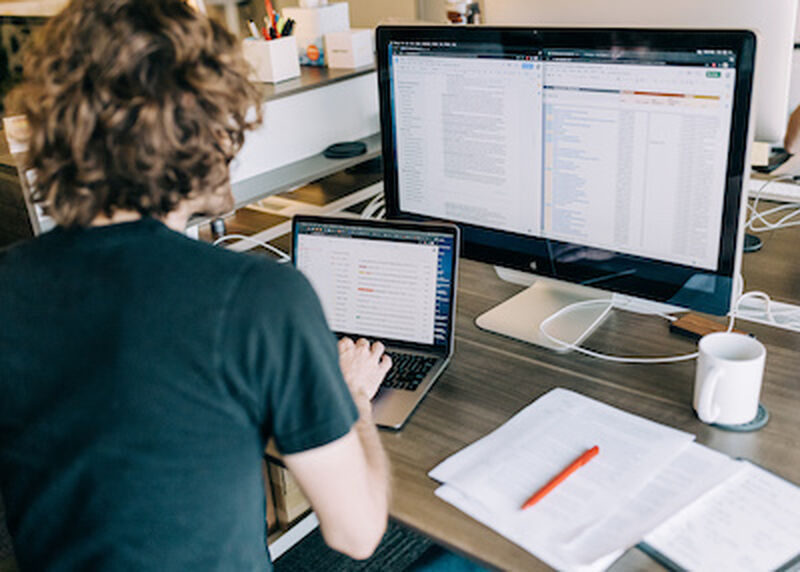 person sitting in front of laptop and larger screen. Red pen on stack of paper to the right.